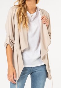 Veste en beige avec un tissu doux, devant drapé et manches retroussées. Portée sur une chemise blanche et associée à un jean bleu.