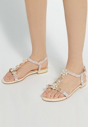 Platte sandalen met een doorzichtige band versierd met bloemige versieringen in crème en gouden accenten. Een gladde, lichtgekleurde zool.