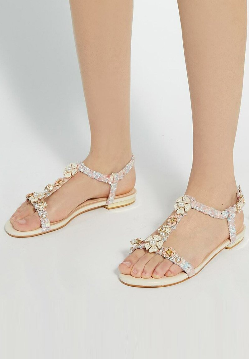Flat sandals with a translucent strap adorned with floral embellishments in cream and gold accents. A smooth, light-colored sole.