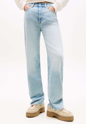 Person iført lyseblå jeans med vidde i benene, med hænderne i lommerne og beige loafers med tyk sål på hvid baggrund.