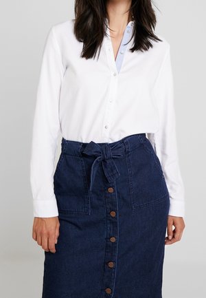 Frau trägt ein weißes Hemd mit Knöpfen, das in einen hoch taillierten, dunklen Jeansrock mit vorderen Knöpfen und einem gebundenen Stoffgürtel gesteckt ist.