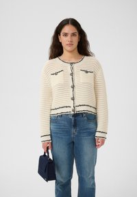 Kvinna som bär en vit texturerad kofta med svart kantband, blå jeans, håller en liten mörkblå handväska och står mot en enfärgad bakgrund.