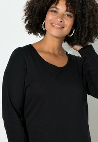 Zwarte top met lange mouwen en een ronde halslijn, met een zachte textuur. De drager heeft krullend haar en raakt met één hand de nek aan.