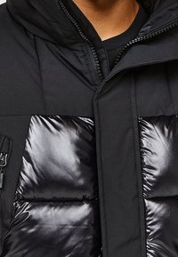 Svart pufferjacka med glänsande quiltade sektioner och matt yta. Huva med dragkedja fram och två sidofickor.