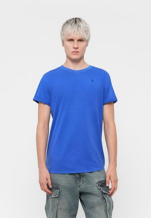 Tricou albastru cu guler rotund și mâneci scurte răsucite, realizat dintr-un material moale, cu un detaliu discret al logo-ului pe piept, asortat cu pantaloni cargo gri.