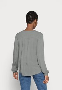Blouse à manches longues en gris clair, confectionnée dans un tissu doux. Caractérisée par un empiècement dos et des plis, avec un ourlet arrondi et une coupe décontractée. Portée avec un jean bleu.