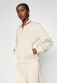 Krämfärgad zip-up-jacka av mjukt material med hög krage, ribbstickade muddar och diskret branding på bröstet. Avslappnad passform med fickor.