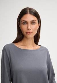 Šedý dlouhý rukáv s lodní výstřihem, vyrobený z měkké tkaniny. Má hladkou texturu a volný střih.