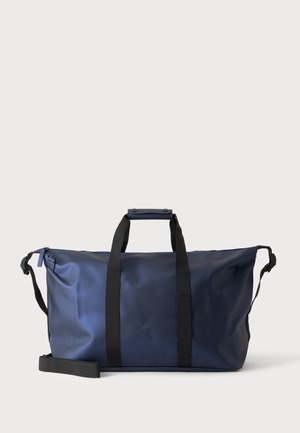 Μεγάλη σκούρη μπλε τσάντα duffel με μαύρες λαβές και ρυθμιζόμενο ιμάντα ώμου σε απλό ανοιχτό φόντο.