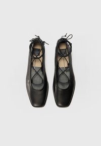 Sapatos de couro pretos com bico quadrado, lados abertos e detalhes de atacadores cruzados. Apresentam uma textura suave e um design minimalista.