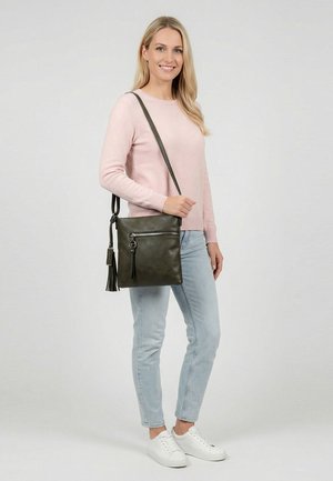 Sieviete, kas valkā gaiši rozā džemperi un gaiši zilas džinsas, tur tumši zaļu plecu somu ar pušķi, stāvot pret vienveidīgu fonu.