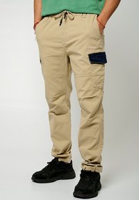 Beige cargobroeken met meerdere zakken, waaronder een donkerblauwe opgestikte zak. Met een trekkoord in de taille en tapered pijpen. Gemaakt met zwarte sneakers.