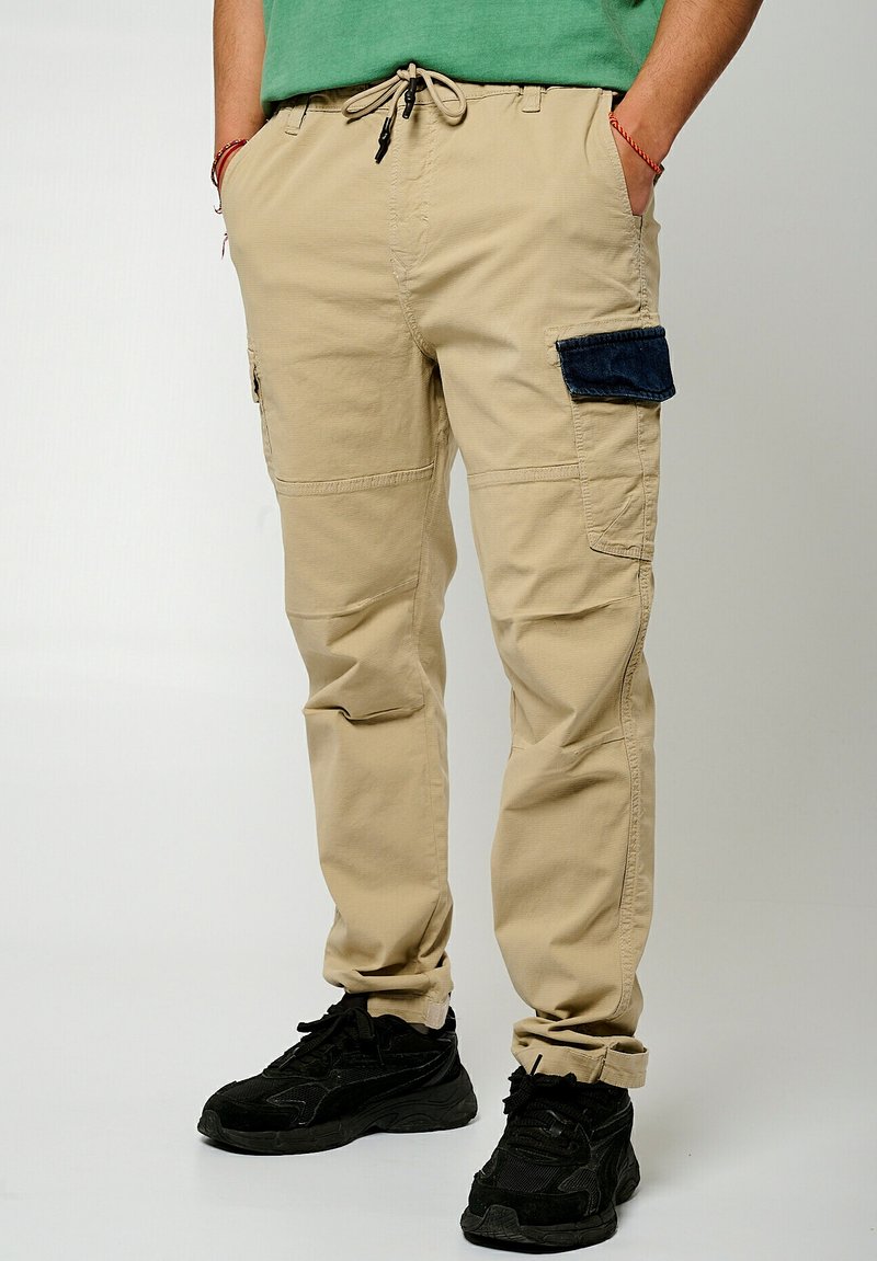 Beige cargobroeken met meerdere zakken, waaronder een donkerblauwe opgestikte zak. Met een trekkoord in de taille en tapered pijpen. Gemaakt met zwarte sneakers.