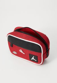 Rote Kosmetiktasche aus Stoff mit einem schwarzen Lederpanel, weißen Akzenten und silbernen Details, die Sneaker-Logos zeigen. Mit einem Reißverschluss oben.
