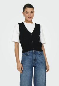 Schwarze Strickweste mit V-Ausschnitt und fünf Knöpfen, getragen über einem weißen T-Shirt, kombiniert mit hoch sitzenden blauen Jeans.