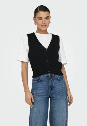 Chaleco de punto negro con escote en V y cinco botones, llevado sobre una camiseta blanca, combinado con unos vaqueros de talle alto en azul.