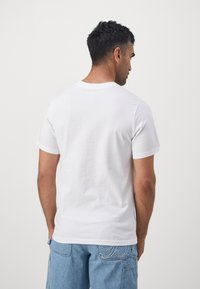 Weißes Baumwoll-T-Shirt mit Rundkragen und kurzen Ärmeln, das eine glatte Textur aufweist. Getragen mit hellblauen Jeansshorts.