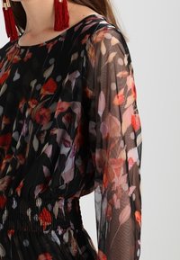 Průsvitné černé šaty s květinovým vzorem v červené a růžové barvě, dlouhými rukávy, stahovacím pasem a puntíky. Doplněné náušnicemi s třásněmi.