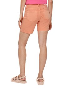 Różowe spodenki dżinsowe w kolorze brzoskwiniowym o dopasowanej talii, z tylnymi kieszeniami i końcu powyżej kolana. Nosi je z jasnobrązowymi sandałami na paski.