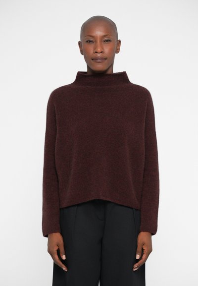 Bordowy, oversize sweter z wysokim kołnierzem i długimi rękawami, wykonany z miękkiego, teksturowanego materiału. Nosi się go na czarnych spodniach.