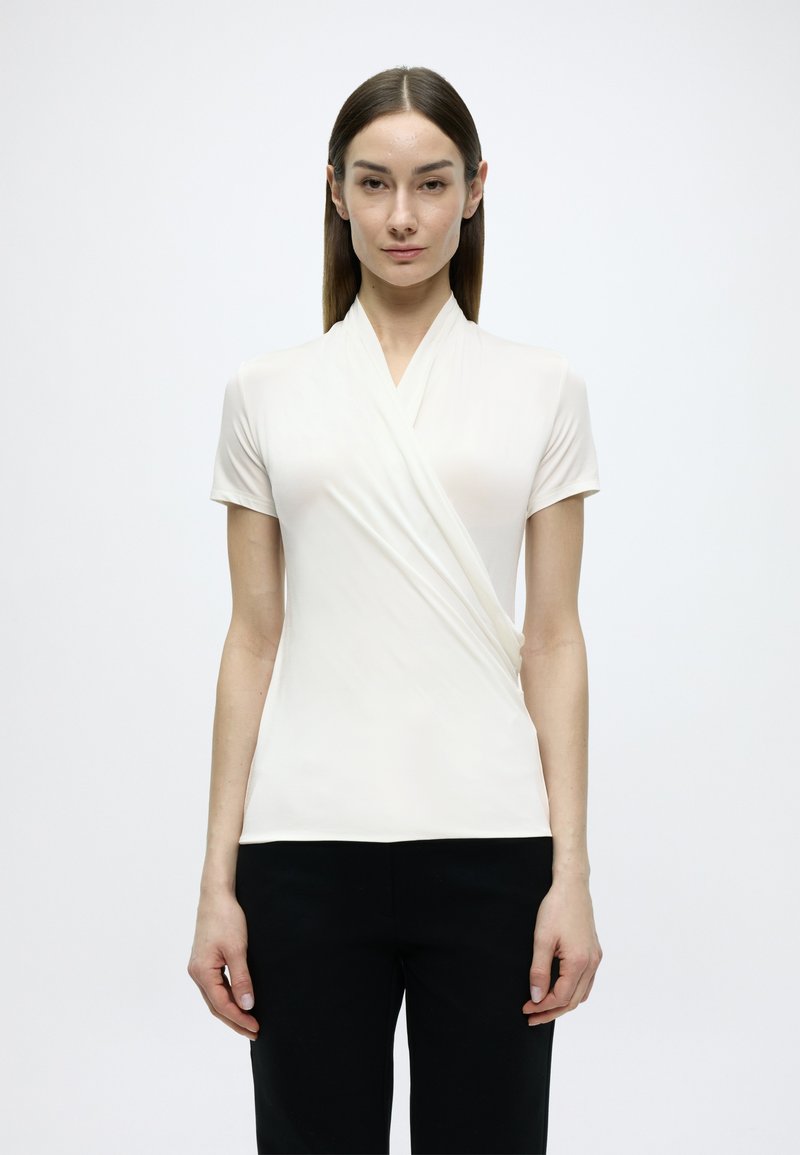 Κοντομάνικη μπλούζα τύπου wrap σε απαλό κρεμ ύφασμα με σχεδιασμό V-neck, που διαθέτει λεία υφή και εφαρμοστή σιλουέτα. Συνδυάζεται με μαύρο παντελόνι.