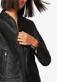 Mustat nahkatakki, jossa on etuvetoketju, uurrettu kaulus ja kaksi sivutaskua vetoketjulla. Teknologia on sileä ja istuva malli.