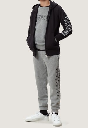 Person iført grå grafisk sweatshirt og joggingbukser med sort lynlåstrøje, hænder i lommerne og hvide sneakers foran ensartet baggrund.