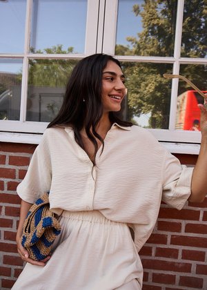 Vrouw glimlacht buiten bij een bakstenen muur en raam, draagt een losse crème outfit en houdt een blauw en beige gevlochten tas vast.