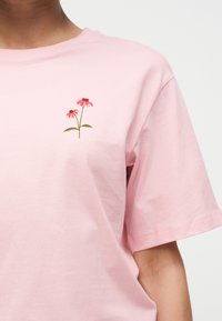 Άτομο που φοράει ανοιχτό ροζ μπλουζάκι με μικρό κεντητό σχέδιο δύο ροζ κωνοφόρων στο στήθος.