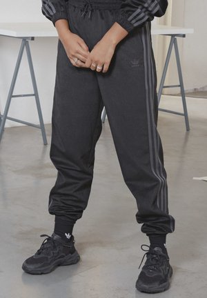 Osoba stoi w pomieszczeniu, ubrana w czarne dresowe spodnie Adidas, czarne skarpetki, czarne sportowe buty, z rękami splecionymi przed ciałem.