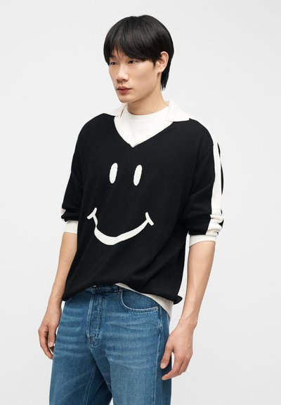 Νεαρός άνδρας με μαύρα μαλλιά, φοράει μαύρο πουλόβερ με μεγάλο λευκό χαμογελαστό πρόσωπο και μπλε τζιν, στέκεται μπροστά σε απλό φόντο.