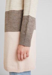 Garnit kofta med randig design i kräm, taupe och ljusrosa färger, ribbade fållar och en mjuk textur.