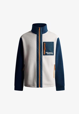 Balts un tumši zilas krāsas flease jaka ar augstu apkakli, oranžu rāvējslēdzēju un krūšu kabatu ar rāvējslēdzēju un "RED BULL RACING" logotipu.