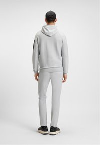 Férfi világosszürke kapucnis pulóverben, hozzá illő nadrágban, fekete sapkában és fekete sportcipőben áll, háttal a sima háttérnek.