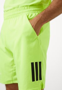 Pantalones cortos deportivos de color verde neón con cinturilla elástica, bolsillo lateral y tres rayas verticales negras en la pierna izquierda. Textura de tela suave.