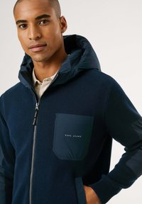 Hombre con una sudadera con cremallera azul marino, capucha acolchada y bolsillo en el pecho etiquetado "PEPE JEANS," mano en el bolsillo lateral, fondo neutro.