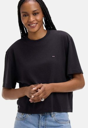 Femme aux longs cheveux tressés souriante, portant un t-shirt noir ample et un jean bleu clair, les mains jointes devant elle.