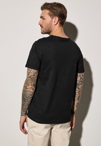 Camiseta negra de manga corta con cuello redondo, fabricada en un material ligero y con un corte holgado, wear por una persona que muestra intrincados tatuajes en sus brazos.
