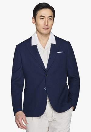 Uomo con blazer blu navy sopra una camicia bianca con colletto e pantaloni chiari, in piedi con una mano in tasca contro uno sfondo neutro.