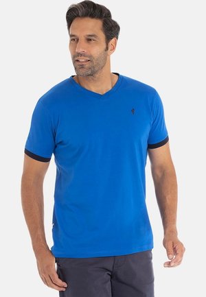 Homme avec des cheveux courts et foncés portant un t-shirt bleu à col en V avec une bordure sombre sur les manches et un pantalon foncé, regardant légèrement vers la gauche.