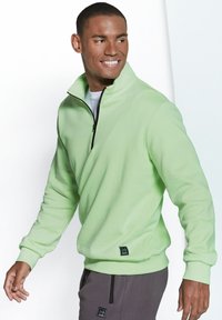Light green fleece pullover with a half-zip collar, ribbed cuffs, and hem. Features a small black logo patch near the waistband.