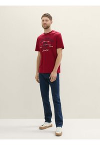 Rød bomulds t-shirt med korte ærmer, med sort trykt logo og tekst. Kombineret med mørkeblå jeans og hvide sneakers med beige såler.