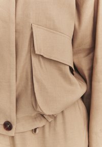 Gros plan d'une poche de veste en tissu beige avec rabat, bouton visible et couture détaillée sur un matériau texturé.