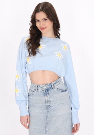 Ung kvinde med langt brunt hår iført en lyseblå kort sweater med hvide og gule blomster samt en højttaljet denimskørt.