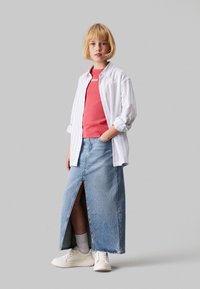 Falda maxi de mezclilla con una abertura frontal, combinada con una blusa de coral de canalé y una camisa blanca a rayas con botones. Zapatillas blancas y calcetines cortos completan el look.