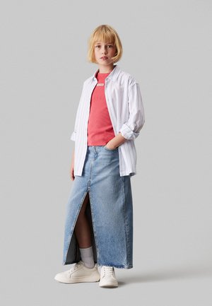 Gonna lunga in denim con spacco frontale, abbinata a un top a coste corallo e una camicia bianca a righe con bottoni. Sneakers bianche e calzini alla caviglia completano il look.