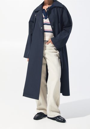 Personne portant un long manteau bleu marine sur un pull en maille blanc avec des rayures bleues et rouges, un pantalon beige à jambes larges et des mocassins noirs.