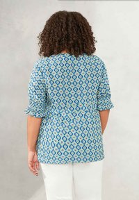 Blusa blu a fantasia con maniche corte a volant, decorata con motivi floreali e a diamante in bianco e verde, indossata con pantaloni bianchi.
