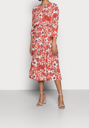 Femme portant une robe midi corail avec un motif floral blanc et rose et des manches trois-quarts, assortie à des sandales beiges à talons hauts.