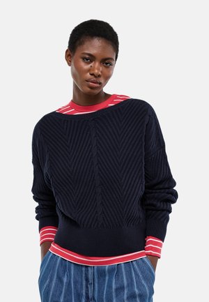 Femme portant un pull texturé bleu marine par-dessus une chemise à rayures rouges et blanches, avec un pantalon à fines rayures bleues, debout devant un fond blanc.